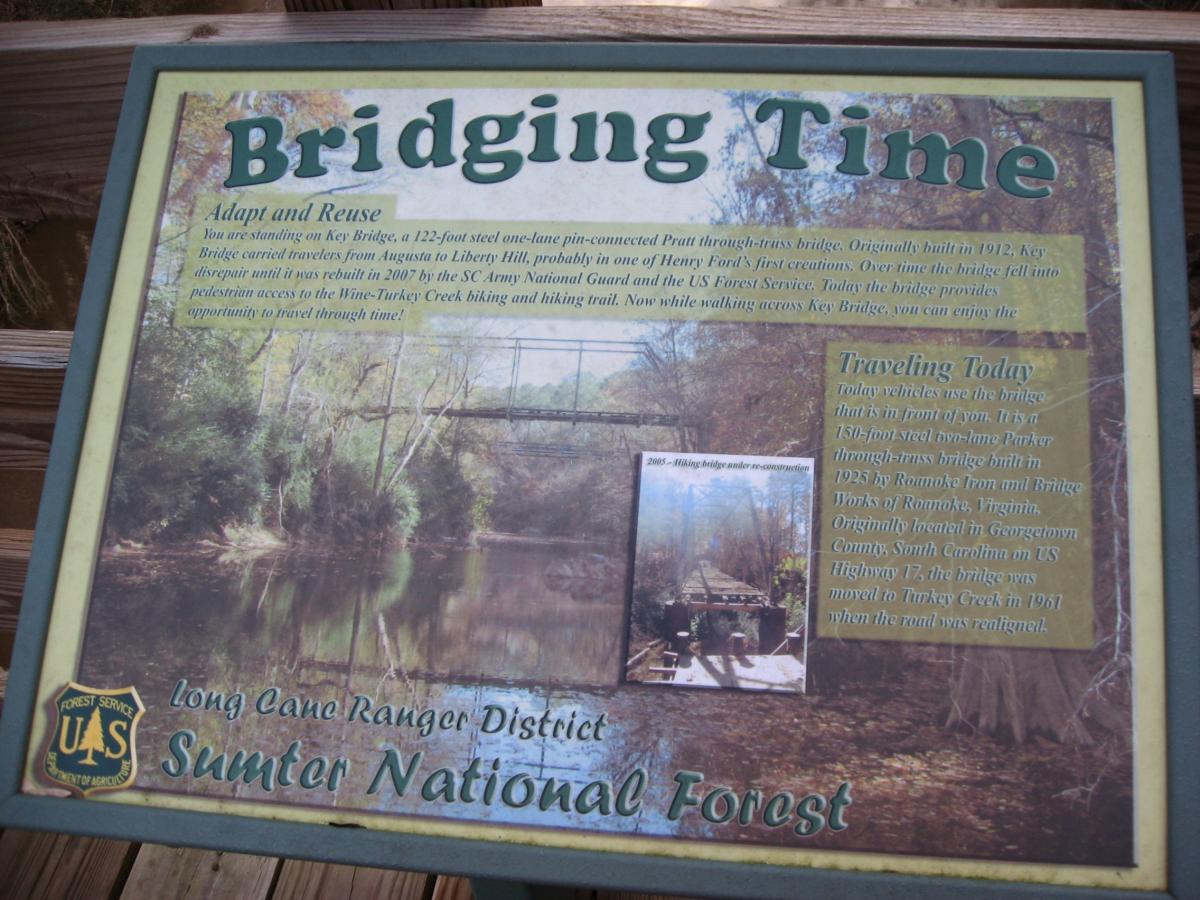 Image of a informational sign titled "Bridging Time" located in the Sumter National Forest. The sign describes the Key Bridge, a 122-foot steel one-lane pin-connected Pratt through-truss bridge, originally built in 1912 and rebuilt in 2007. It provides historical context about the bridge's construction, its use over time, and its connection to the surrounding nature. A smaller photo of the bridge is included on the sign. The sign features the U.S. Forest Service logo in the corner. Turkey Creek mountain bike trail.