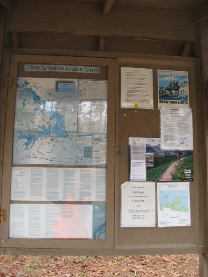 A wooden information board displaying a map of J. Strom Thurmond Dam and Lake at Clark's Hill, along with various posted notices and guides about safety and regulations. The board includes a phone number for emergencies, trail maps, and information related to recreational activities in the area. Wine Creek mountain bike trail.