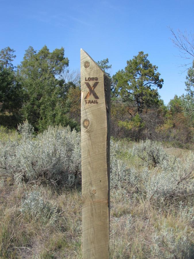Wooden trail marker labeled "Long X Trail" amidst a natural landscape with green trees and shrubs, under a clear blue sky. Long X mountain bike trail.