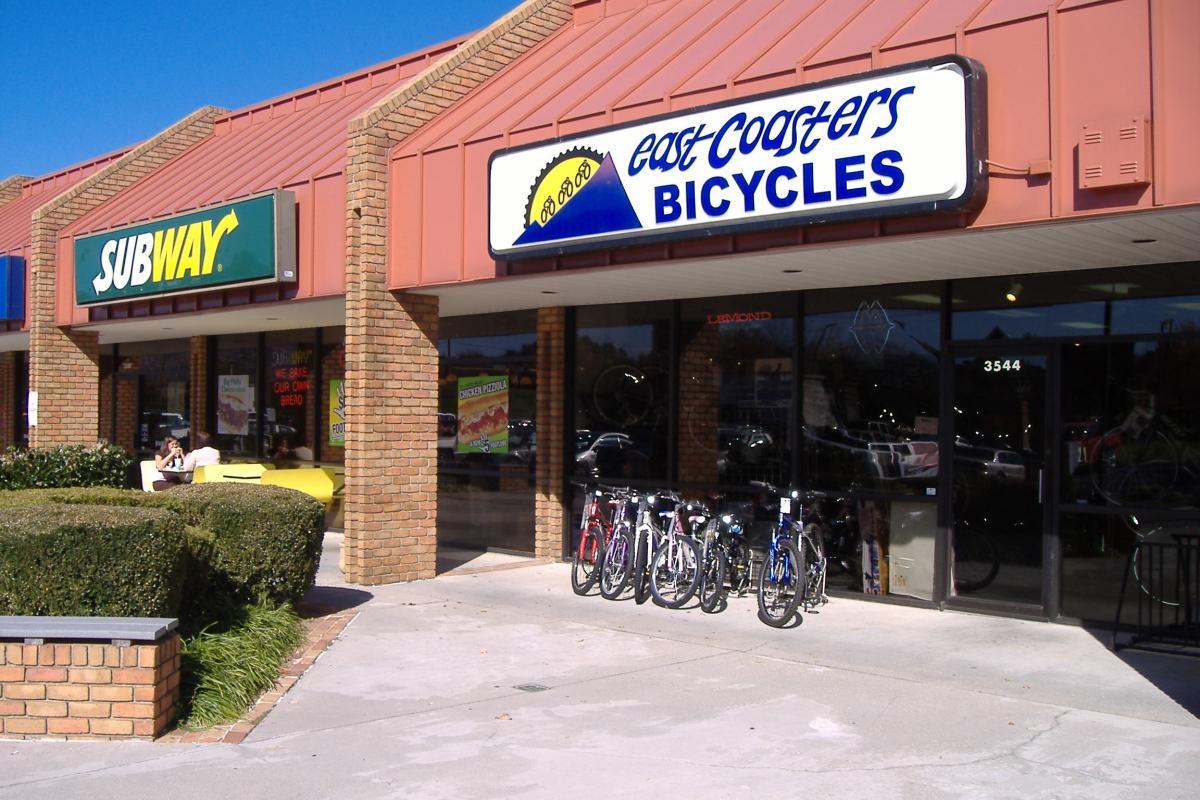Exterior view of a shopping center featuring a Subway restaurant and a bicycle shop named East Coasters Bicycles. The bicycle shop displays several bikes for sale outside, and there is greenery in front of the buildings under a clear blue sky.