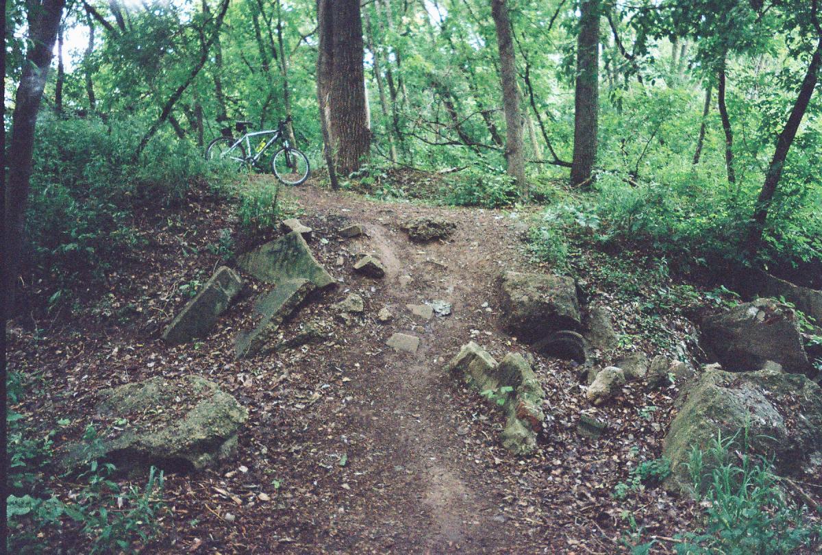 A winding dirt path through a lush green forest, flanked by large rocks and scattered leaves. A bicycle leans against a tree in the background, suggesting an outdoor adventure scene. Baird Creek mountain bike trail.