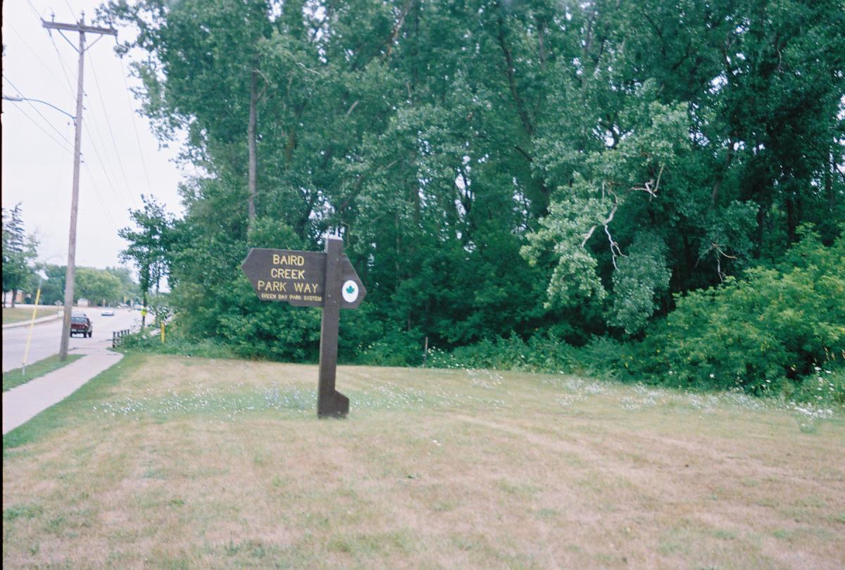 A wooden sign indicating "Baird Creek Park Way" with a green dot symbol, set against a backdrop of trees and grassy land. A road with a car and utility poles is visible in the background. Baird Creek mountain bike trail.