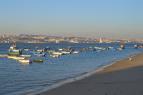 A scenic view of a beach with several small boats floating in calm water, set against a backdrop of distant hills and a clear blue sky. Fonte Da Telha mountain bike trail.