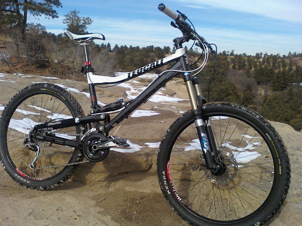 A mountain bike is positioned on a rocky surface with a backdrop of trees and a clear blue sky. The bike features a black and white frame with suspension components and knobby tires, showcasing its design for off-road cycling. Snow can be seen on the ground around the bike, indicating a winter setting. Palmer Park mountain bike trail.