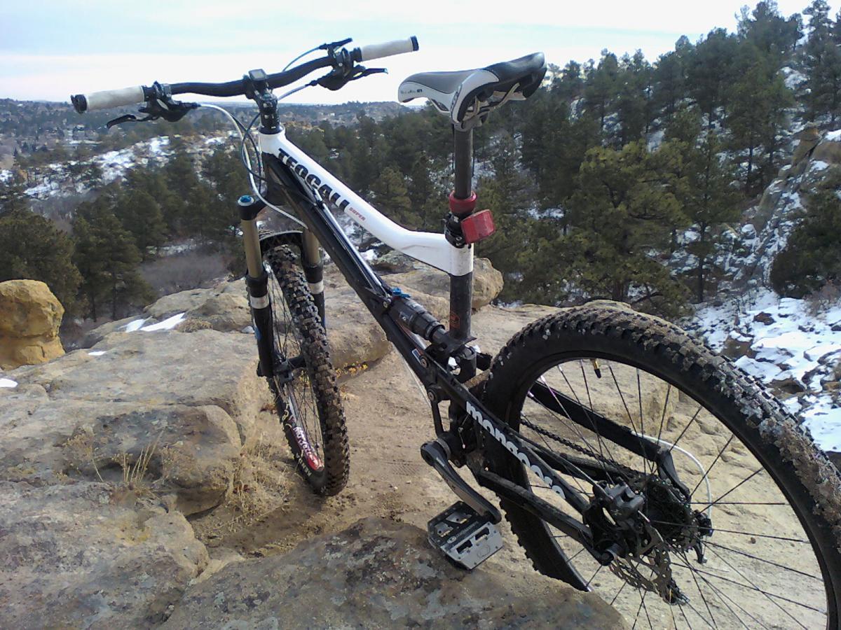 A black and white mountain bike resting on rocky terrain with a scenic view of pine trees and snow in the background. The bike features thick, textured tires and a slightly muddy frame, indicating recent use. Palmer Park mountain bike trail.