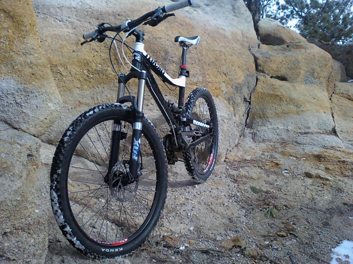 A mountain bike leaning against a rocky surface, with snow on the ground and scattered pine needles nearby. The bike features a black frame, thick tires, and front suspension, suitable for off-road trails. Palmer Park mountain bike trail.