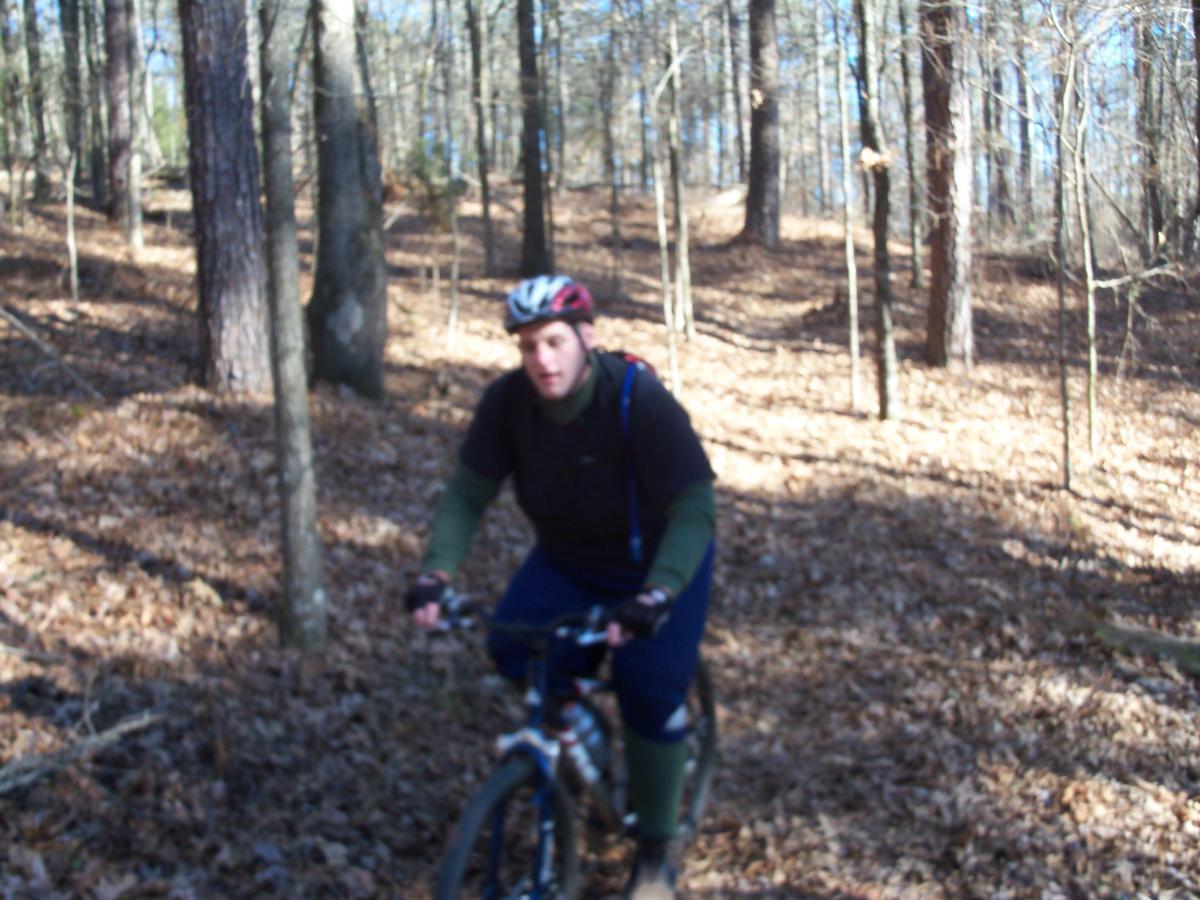 A person riding a mountain bike on a trail in a wooded area during autumn, surrounded by trees and fallen leaves. The rider is wearing a helmet, a dark shirt, and green pants, with a backpack visible on their back. The background features a mix of tree trunks and foliage. Hickory Knob State Park mountain bike trail.