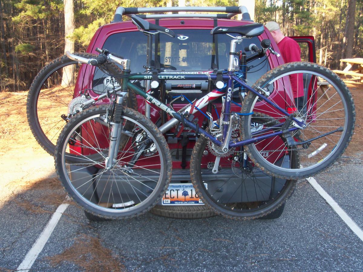 Alt text: Two bicycles are mounted on the back of a red vehicle parked in a forested area. The vehicle's rear window displays a state park sticker, and there is a person partially visible in the background. Hickory Knob State Park mountain bike trail.