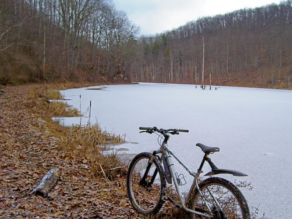 A mountain bike resting on the shore of a frozen pond, surrounded by autumn leaves and bare trees in a wooded area. The water is covered with ice, and the scene reflects a quiet, tranquil winter landscape. North Bend State Park mountain bike trail.