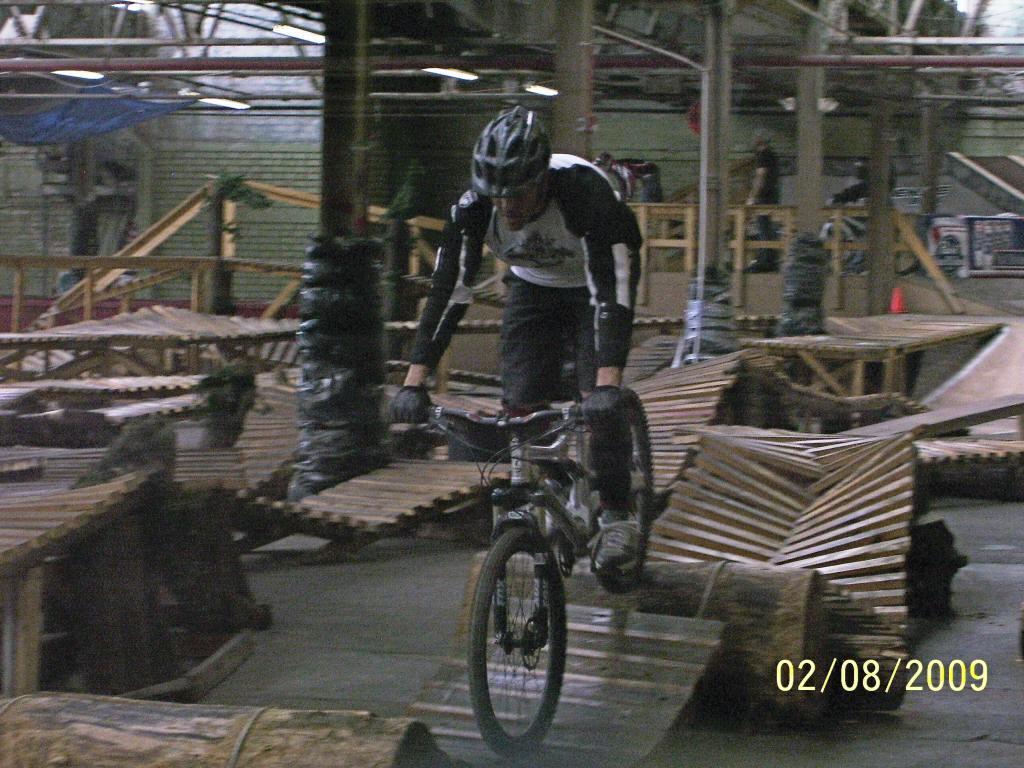 A mountain biker navigating a challenging indoor skate park course, featuring wooden ramps and obstacles. The rider, dressed in a black and white jersey and helmet, is captured in mid-ride on a mountain bike, showcasing skills and balance. Soft lighting and a variety of wooden structures create a dynamic environment. Rays Indoor Mtb Park mountain bike trail.