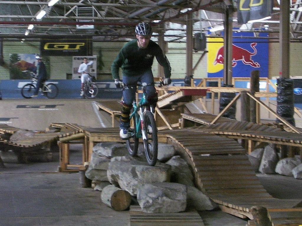 A cyclist performing a trick on a mountain bike over rocks and wooden ramps in an indoor bike park, surrounded by other riders. The environment features a mix of ramps and obstacles designed for biking, with various logos on the walls. Rays Indoor Mtb Park mountain bike trail.