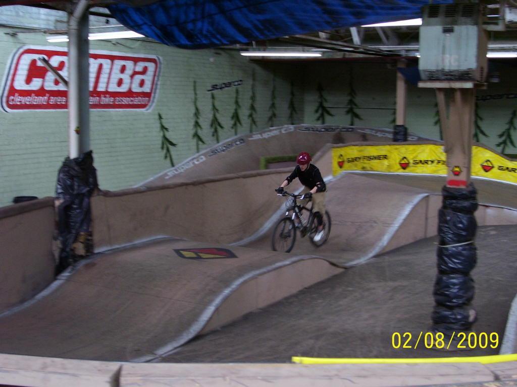 A person riding a bicycle on an indoor pump track with various wave-like features. The background includes colorful banners and a logo for a biking association. The scene is set in a warehouse-like environment with green walls and wooden supports. Rays Indoor Mtb Park mountain bike trail.