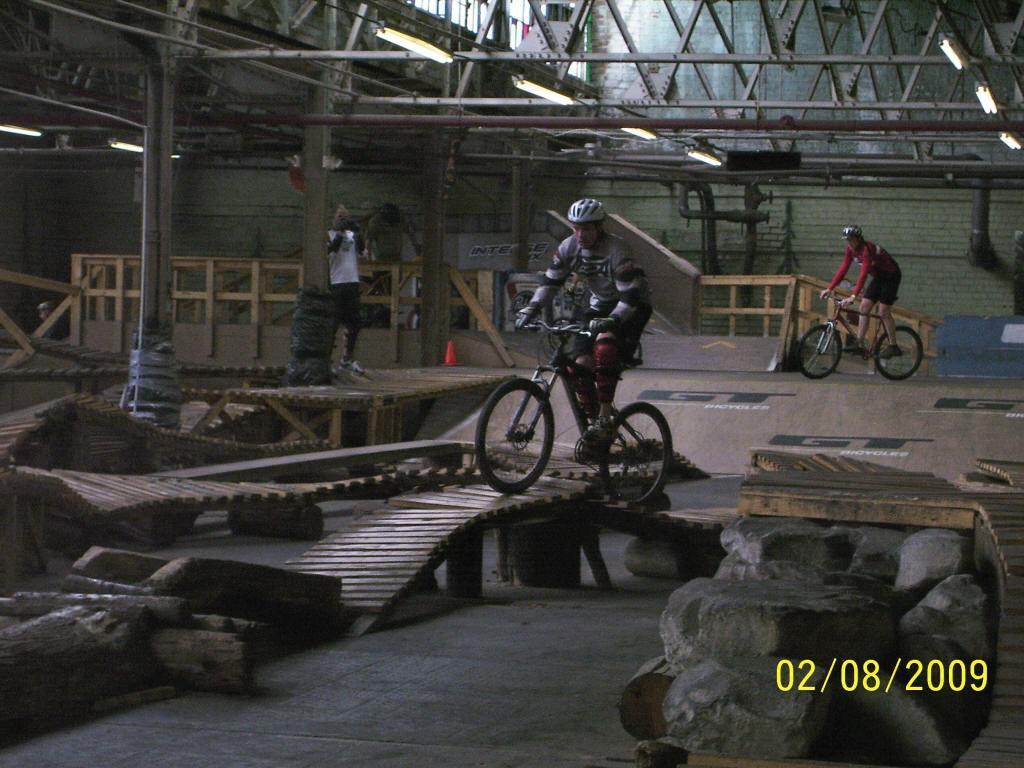 A biker navigates a wooden obstacle course in an indoor biking facility, surrounded by wooden ramps and rock features. Another biker is visible in the background, while a person captures the moment with a camera. The date in the corner reads 02/08/2009. Rays Indoor Mtb Park mountain bike trail.