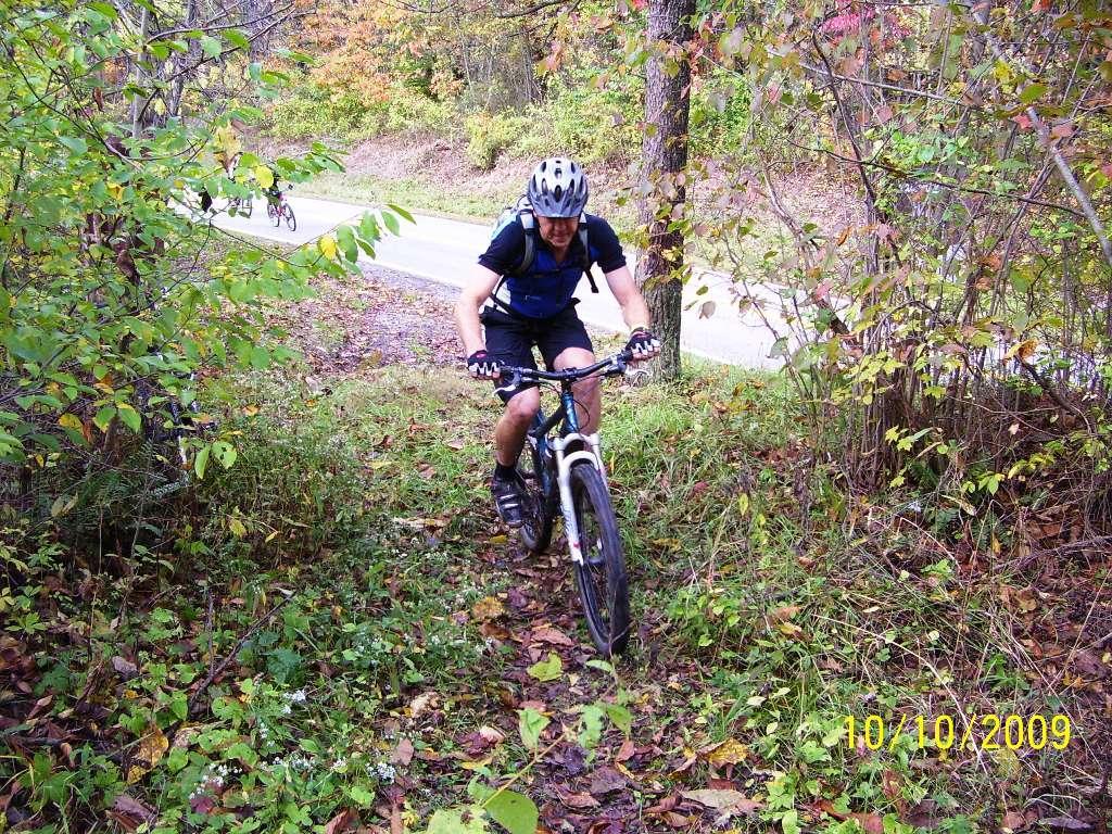 A mountain biker navigating a trail surrounded by greenery and autumn foliage, with a road visible in the background. The cyclist is wearing a helmet and riding a mountain bike, focused on the path ahead. Ohio View Trail mountain bike trail.