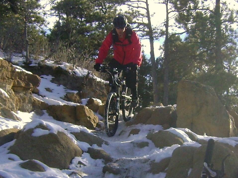A mountain biker in a red jacket navigates rocky terrain covered in snow, surrounded by trees in a winter landscape. Palmer Park mountain bike trail.