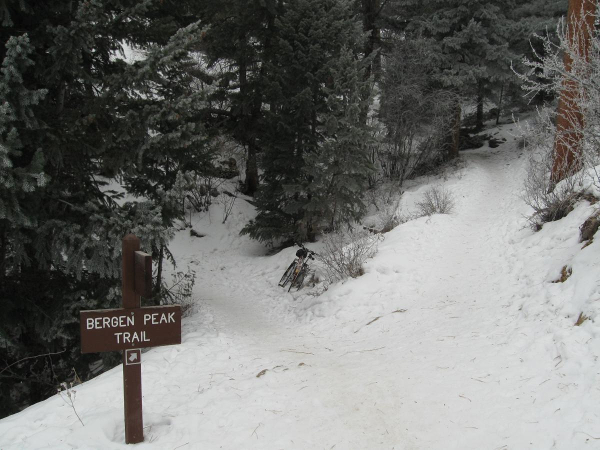 A snow-covered path diverges in a forest, marked by a brown sign that reads "Bergen Peak Trail." Surrounding pine trees are dusted with snow, creating a serene winter landscape. Elk Meadow mountain bike trail.