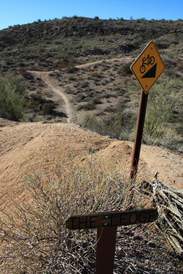 A dirt trail winding through a desert landscape, with a bicycle warning sign indicating a downhill incline. In the foreground, a wooden sign reads "THE LEOGE." The scenery features sparse vegetation and rocky hills under a clear blue sky. McDowell Mountain Park mountain bike trail.