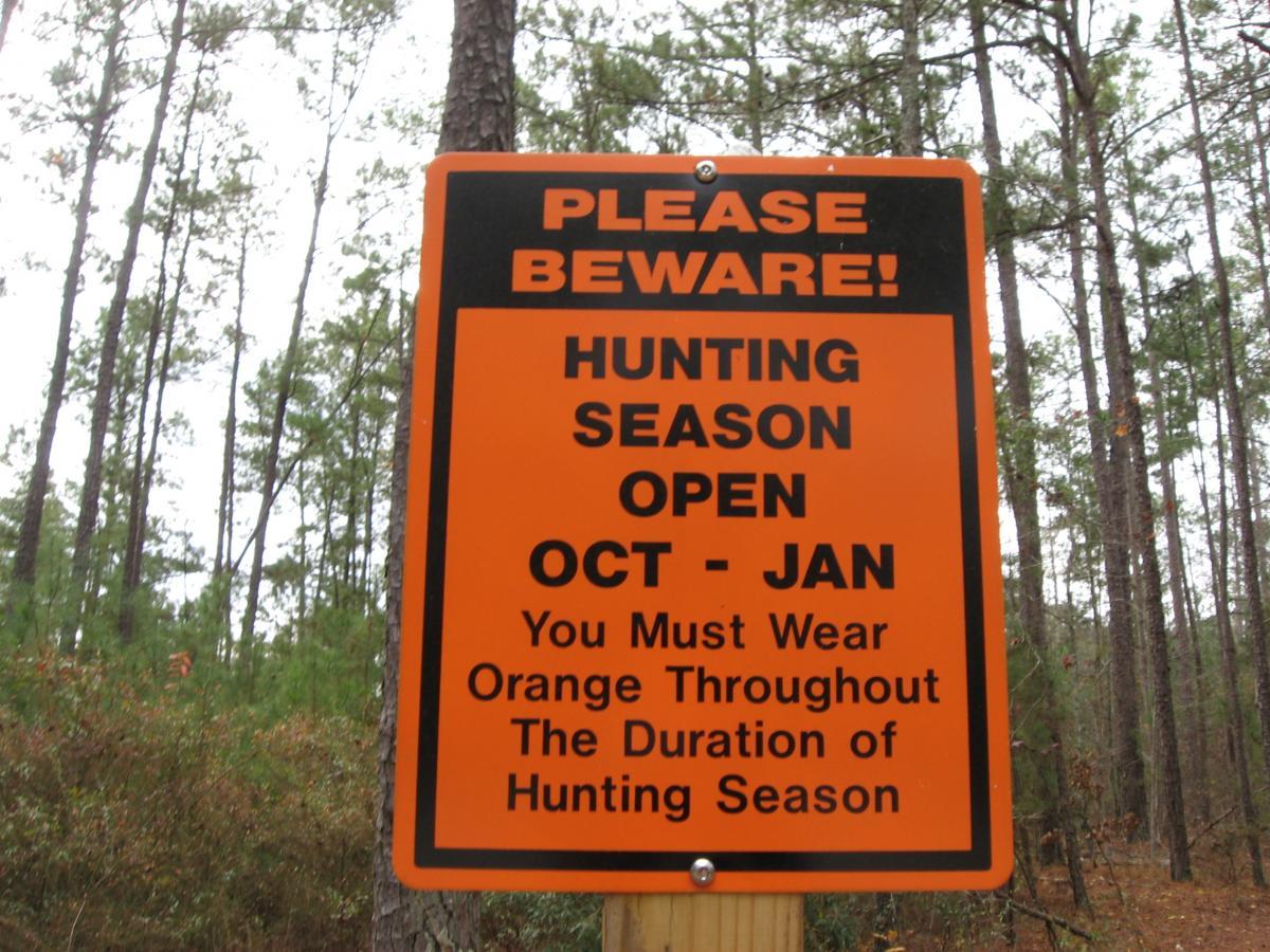 A bright orange warning sign in a wooded area indicating that hunting season is open from October to January. The sign advises individuals to wear orange clothing during this time for safety. Bartram Trail / West Dam / Wildwood Park mountain bike trail.