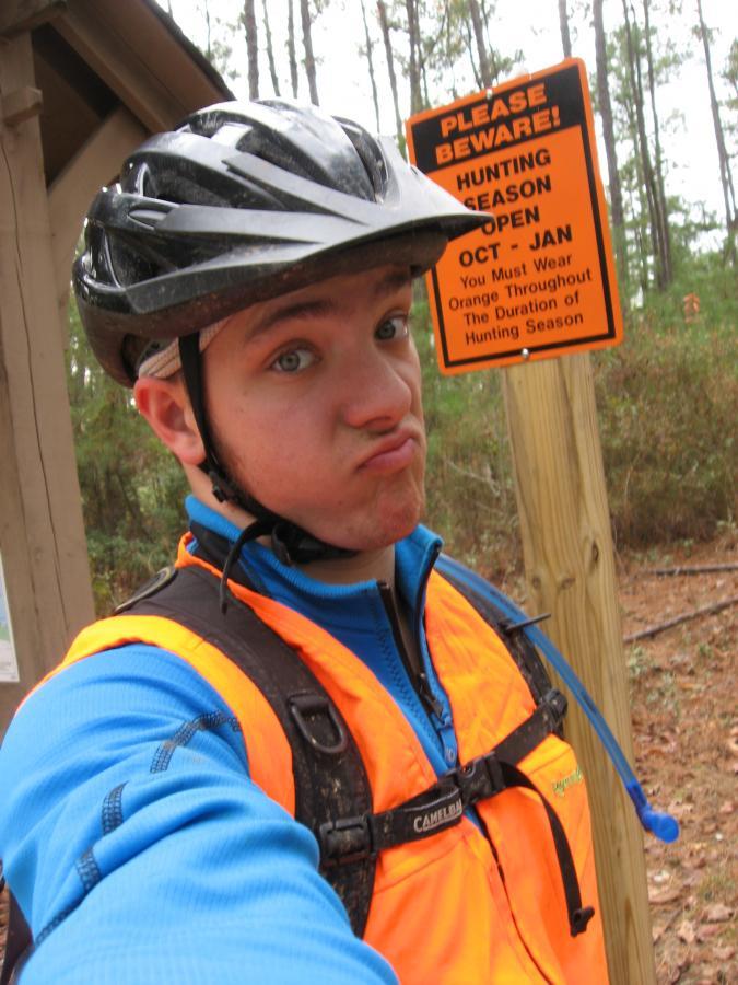A young person wears a bike helmet and an orange safety vest while making a playful face. In the background, there is a warning sign about hunting season, indicating it is open from October to January and advising to wear orange during that time. The setting appears to be a wooded area. Bartram Trail / West Dam / Wildwood Park mountain bike trail.