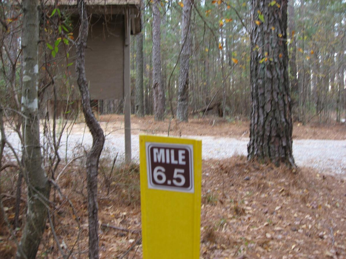A yellow mile marker sign reading "MILE 6.5" stands in a wooded area, with a dirt path visible in the background. Trees surround the marker, and there are fallen leaves on the ground, indicating a natural outdoor setting. A wooden structure is partially shown to the left of the image. Bartram Trail / West Dam / Wildwood Park mountain bike trail.
