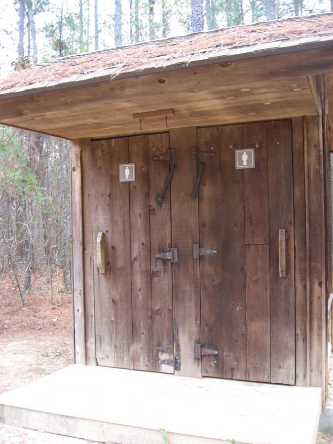 A rustic wooden restroom structure with two doors, each marked with gender signs, situated in a wooded area. The roof is covered with pine needles, and the surrounding environment features trees and pine needles on the ground. Bartram Trail / West Dam / Wildwood Park mountain bike trail.