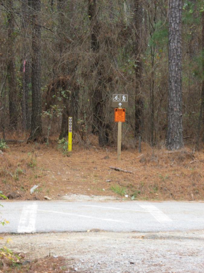 A pathway entry into a wooded area, marked by signage indicating bike and pedestrian access. The signs include icons for bicycles and walking, along with an orange warning notice. The trail is surrounded by trees and pine needles, with a yellow post on the left side displaying a number. The scene is slightly overcast, suggesting a cool day. Bartram Trail / West Dam / Wildwood Park mountain bike trail.