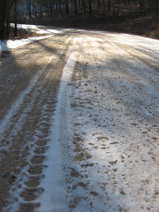 A dirt road partially covered in snow, showing tire tracks and patches of gravel, surrounded by trees. The sunlight casts shadows across the road, creating a contrast between the snow and the dirt. Montgomery Creek Trail mountain bike trail.