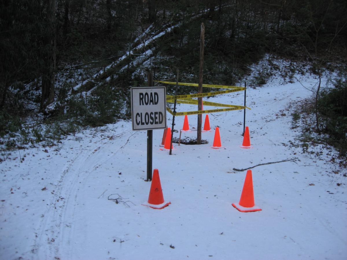 A snow-covered path is blocked by a "ROAD CLOSED" sign and traffic cones, with yellow caution tape marking the closed area. Surrounding trees and underbrush are visible in the background. Montgomery Creek Trail mountain bike trail.