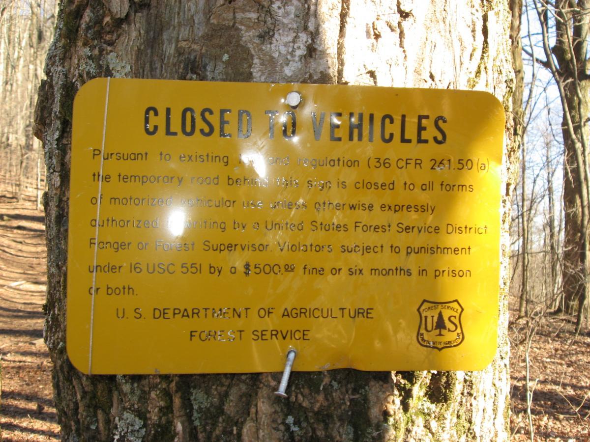 A weathered yellow sign affixed to a tree, displaying the message "CLOSED TO VEHICLES." The sign outlines regulations prohibiting motorized vehicular use on the temporary road, detailing potential penalties for violations. The background features a wooded area with a dirt path, indicating a natural setting. Winding Stairs Loop mountain bike trail.