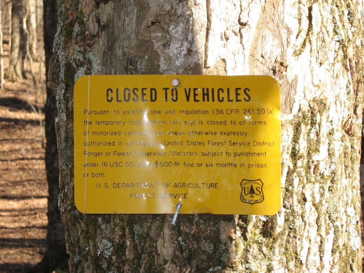 Yellow sign affixed to a tree, reading "CLOSED TO VEHICLES." The sign details that the temporary road is closed to all motorized vehicles, citing legal regulations and potential penalties for violations. The background shows a forested area with trees. Winding Stairs Loop mountain bike trail.