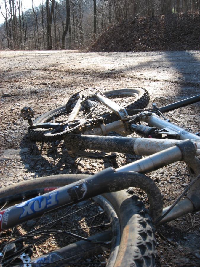 A close-up image of a mountain bike lying on a gravel path, partially obscured by dirt. The surrounding area features trees in the background and a small pile of leaves. Sunlight is casting shadows on the ground, creating a natural outdoor setting. Winding Stairs Loop mountain bike trail.