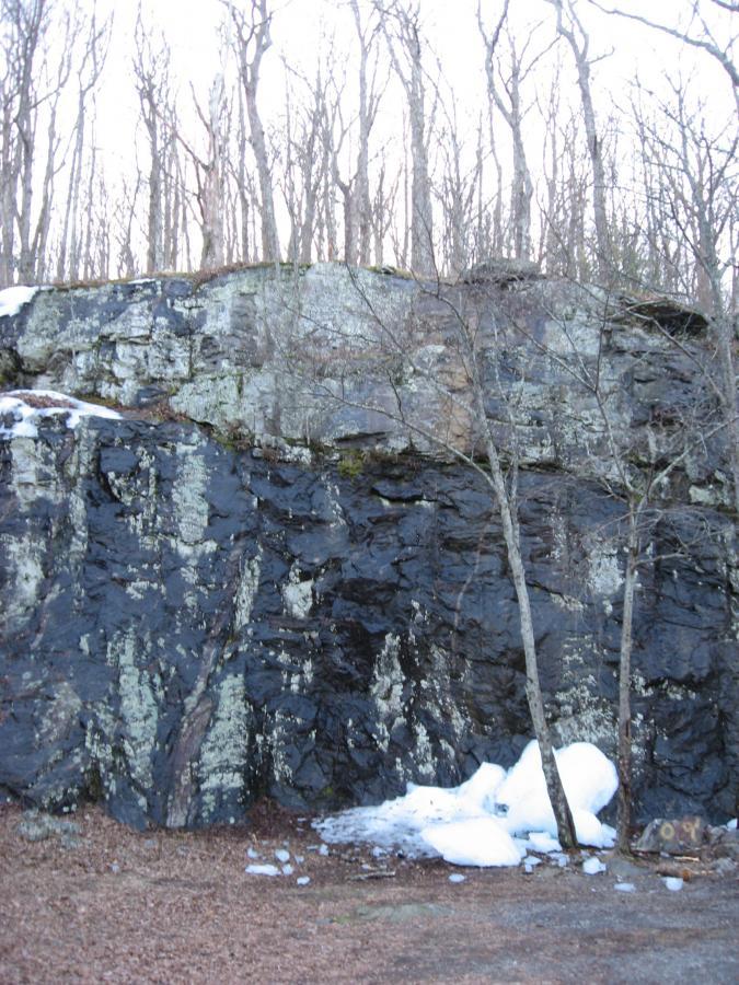 A rocky cliff face with dark, textured stone, partially covered in patches of snow. In the foreground, there are bare trees with thin branches, while the background shows more trees against a pale sky. Some snow and debris are visible on the ground at the base of the cliff. Winding Stairs Loop mountain bike trail.
