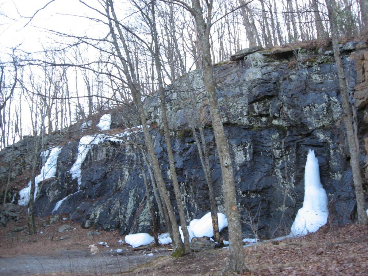 A rocky hillside with exposed dark stones and patches of white snow and ice, surrounded by bare trees. The ground is covered with fallen leaves, indicating early spring or late winter conditions. Winding Stairs Loop mountain bike trail.