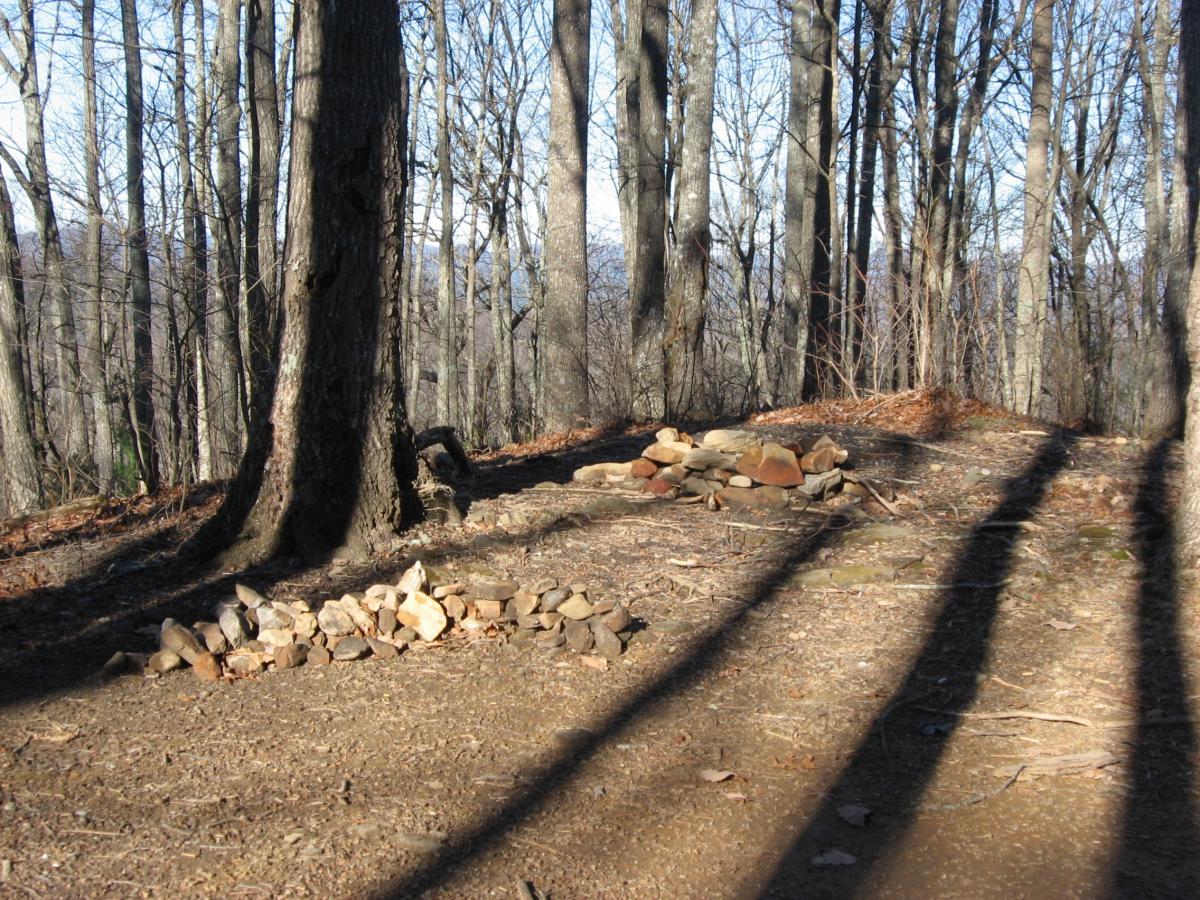 A serene woodland scene featuring a dirt path surrounded by tall, bare trees under clear blue skies. On the left side, there is a neatly arranged pile of stones, with another slightly larger pile on the right. The ground is covered with dry leaves and scattered rocks, with long shadows stretching across the path from the trees. Winding Stairs Loop mountain bike trail.