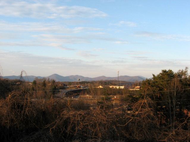A scenic view of distant mountains under a blue sky with scattered clouds, featuring a foreground of shrubbery and trees. In the middle ground, a highway can be seen alongside a small cluster of buildings. The landscape has a tranquil, natural ambiance, indicating a rural or semi-urban setting. Bull / Jake Mountain mountain bike trail.