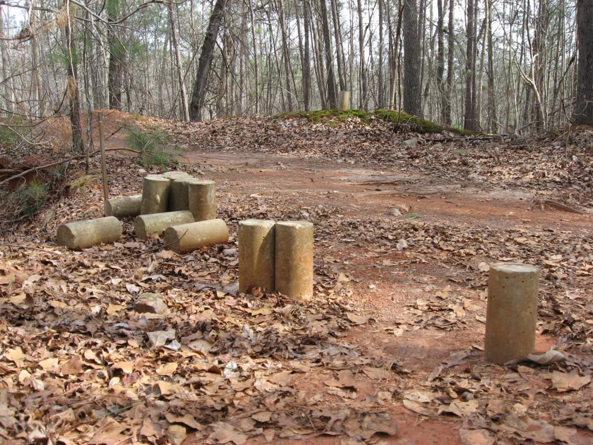 A forest path covered in dry leaves, featuring several concrete or stone cylindrical objects arranged sporadically along the ground. In the background, more trees can be seen, with a slight incline leading to a higher area covered in moss. Chicopee Woods mountain bike trail.