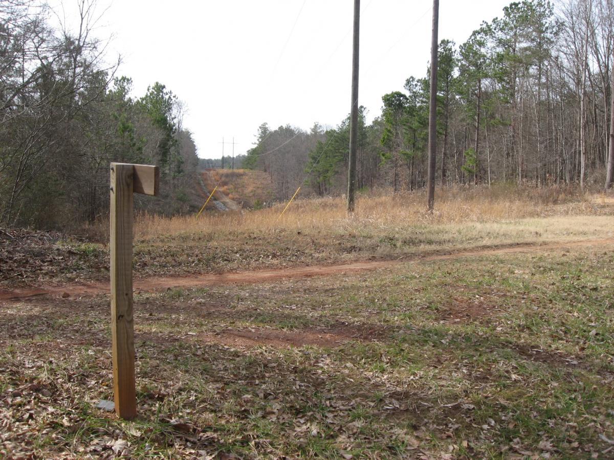 A wooden post stands at the edge of a dirt path, surrounded by sparse grass and scattered leaves. In the background, tall trees line the sides of the path, and utility poles run along a clearing that stretches into the distance. The scene is set in a naturally wooded area, with a mix of brown earth and seasonal foliage. Chicopee Woods mountain bike trail.