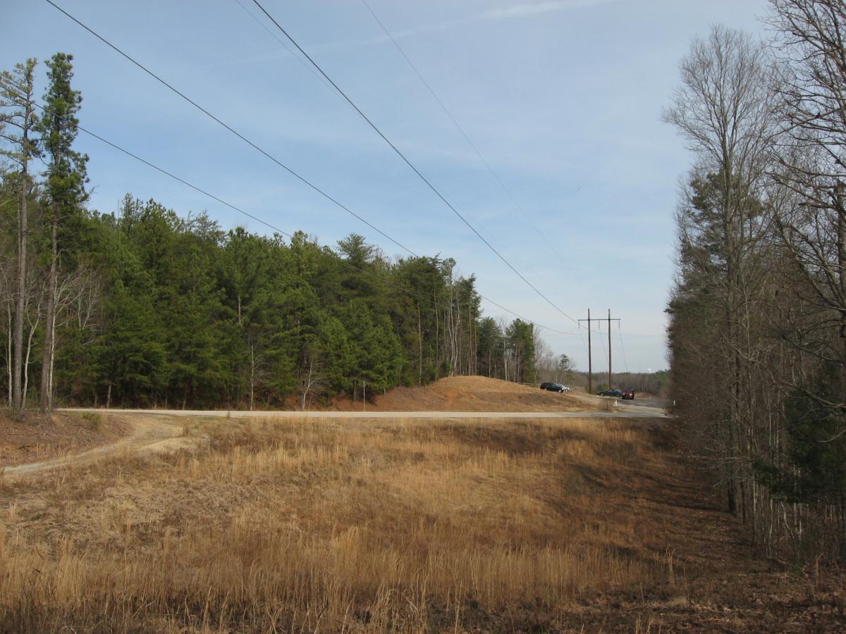 A rural landscape featuring a gravel road that curves into a dirt path, bordered by grassy fields and trees. Power lines run alongside the road, with several vehicles visible in the background near the intersection of the gravel path and a paved road. Overcast skies provide a muted light over the scene. Chicopee Woods mountain bike trail.