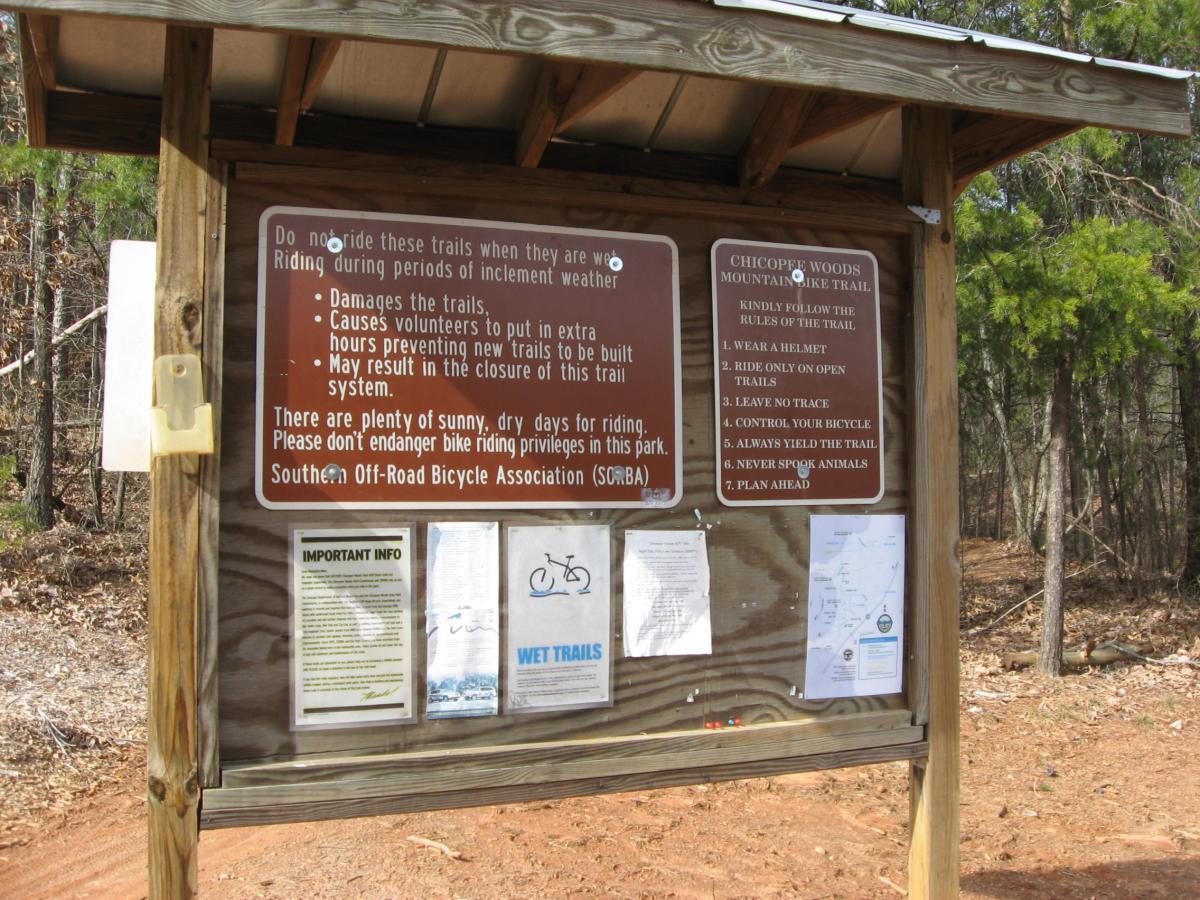 A wooden informational sign at a trailhead, providing guidelines for mountain biking at Chicopee Woods. The sign includes warnings about riding during inclement weather, a list of trail rules, and a reminder to respect the environment. Nearby notices are attached, including maps and additional information for cyclists. Surrounding vegetation suggests a natural setting for outdoor activities. Chicopee Woods mountain bike trail.