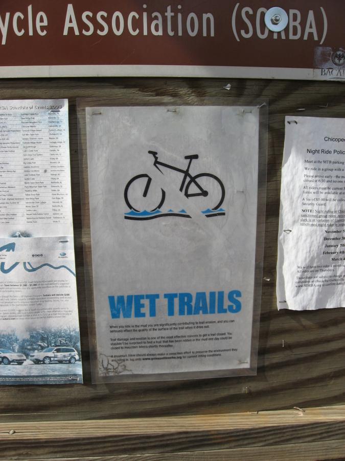 Sign indicating "WET TRAILS" with a bicycle symbol partially submerged in water, mounted on a wooden bulletin board. The sign warns cyclists about the impact of riding on wet trails and encourages responsible trail use. Nearby, there are additional notices and schedules related to the Southern Off-Road Bicycle Association (SORBA). Chicopee Woods mountain bike trail.