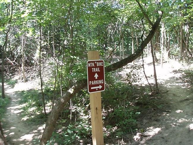 A wooden sign at a fork in the trail, indicating directions for "MTN. BIKE TRAIL" and "PARKING," surrounded by dense greenery and trees in a forested area. Mitchell Memorial Forest Mountain Bike Trail mountain bike trail.
