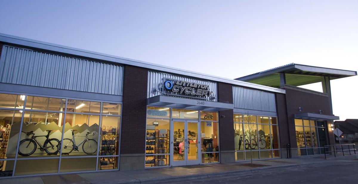 Alt text: Exterior view of Littleton Cyclery, showcasing large glass windows featuring bicycles and outdoor gear. The building has a modern design with metal and brick elements, illuminated in the evening light.