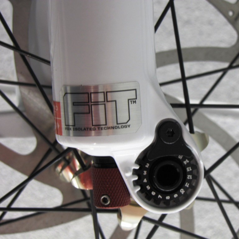 Close-up image of a bicycle fork featuring a label for "FOX ISOLATED TECHNOLOGY" and a dial for adjusting settings. The fork is predominantly white, with a red adjustment knob and visible wheel spokes in the background.