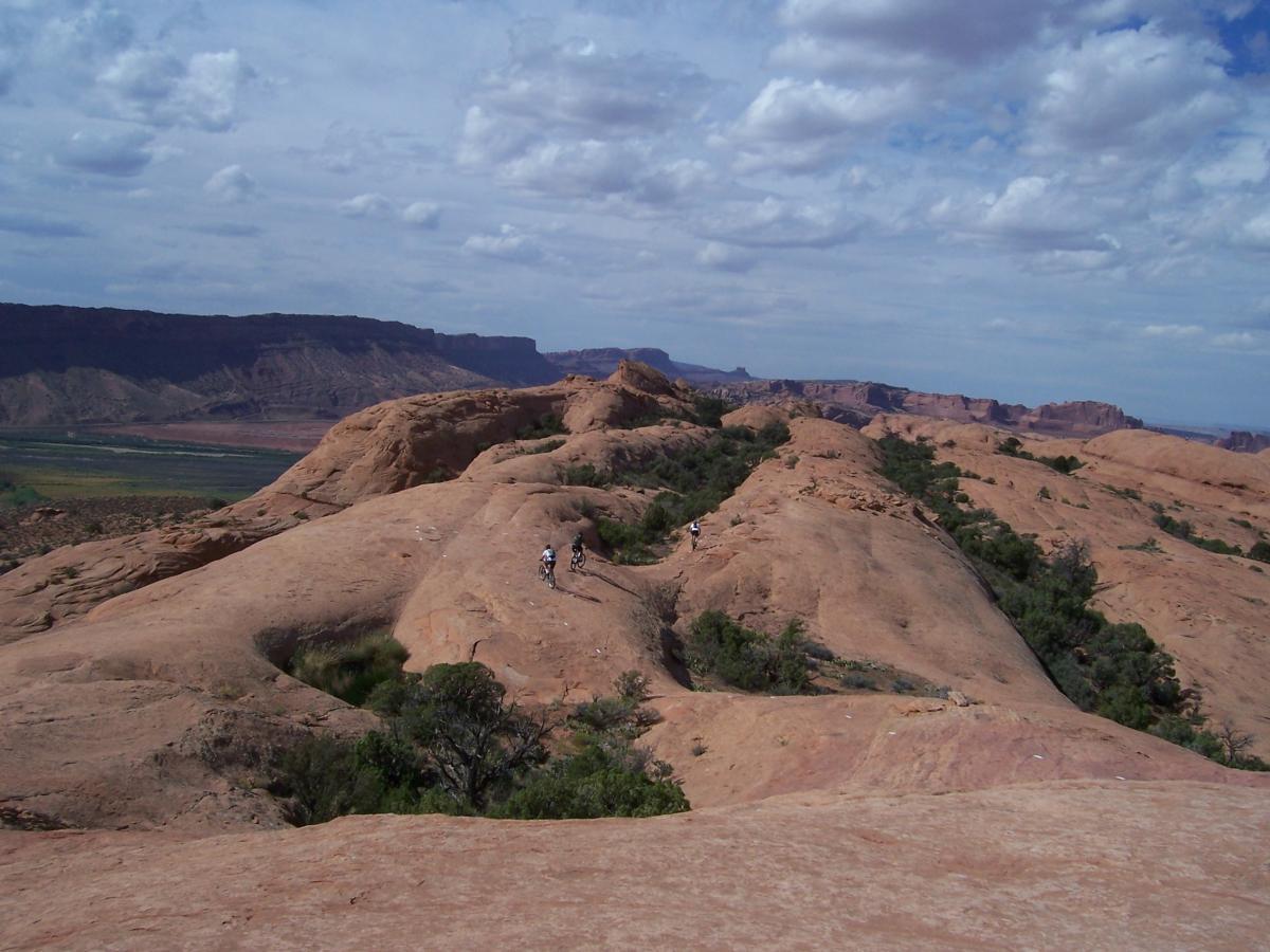 A panoramic view of rugged desert terrain featuring smooth, rocky hills and scattered green vegetation under a partly cloudy sky. A couple of cyclists are riding along a winding path, surrounded by vast canyons and distant cliffs in the background. Slickrock mountain bike trail.
