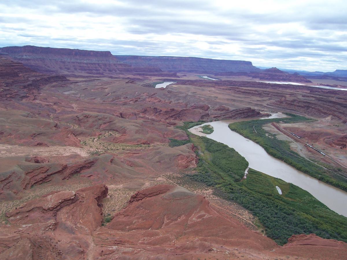Aerial view of a rugged, arid landscape featuring layered red rock formations, with a winding river bordered by lush green vegetation. The sky is overcast, and distant mountains are visible in the background. Amasa Back Trail mountain bike trail.