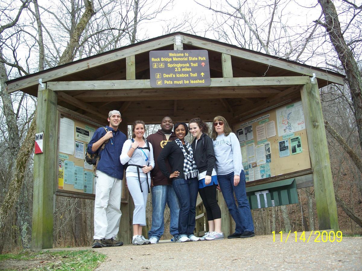 A group of six people stands together in front of a wooden shelter at Rock Bridge Memorial State Park, with signs indicating nearby trails. The scene is set in a natural, wooded area. The group consists of a man and a woman on the left, both wearing outdoor attire, followed by a man in a dark hoodie, a woman in a black and white striped shirt, another woman in a blue shirt, and a woman on the far right in a gray sweater. The date displayed is November 14, 2009. Rock Bridge Memorial State Park mountain bike trail.