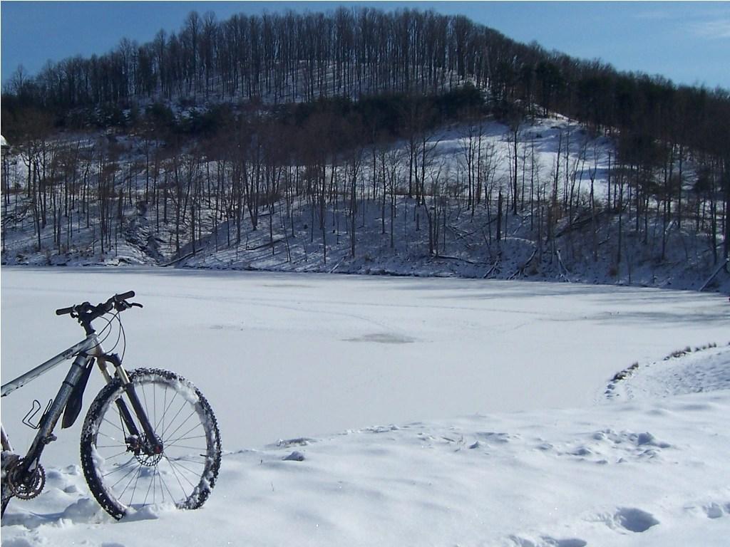 A mountain bike resting on snow-covered ground beside a frozen lake, with a wooded hillside in the background under a clear blue sky. Mountwood mountain bike trail.