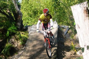 A mountain biker in a yellow and black jersey rides down a dirt ramp in a forested area, surrounded by green foliage and trees. The biker wears a red helmet and rides a red bike, showcasing a dynamic action shot. Amelia Earhart Park mountain bike trail.