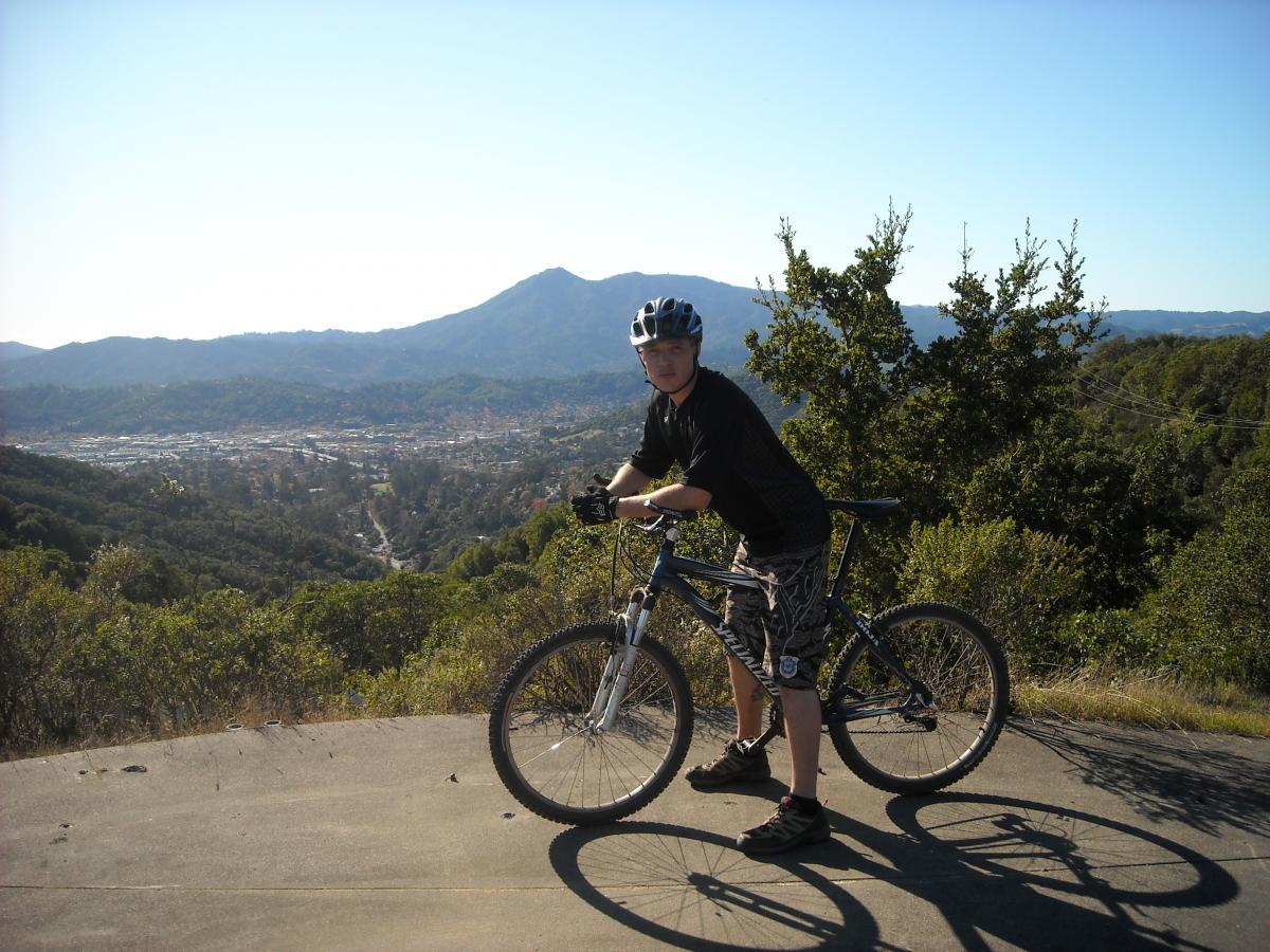 A person standing next to a mountain bike on a scenic overlook, with mountains and a valley in the background under clear blue skies. The individual is wearing a helmet and casual outdoor clothing, looking towards the camera. China Camp mountain bike trail.
