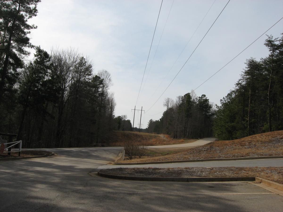 A rural road intersection surrounded by trees and power lines, with a clear sky above. One road veers to the left while another continues straight, with grassy and rocky terrain on either side. Chicopee Woods mountain bike trail.